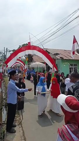 The video captures a street procession, likely a cultural or celebratory event, with a group of people walking down a narrow road. The road is decorated with red and white arches, possibly representing national flags, suggesting a patriotic theme. People are dressed in traditional attire, including women wearing hijabs in various colors like blue and red, and men in shirts and caps. Some individuals are holding flags and parasols. In the foreground, a man in a light blue shirt and black pants is interacting with someone. The environment appears to be a residential area with houses and utility poles visible in the background. The overall atmosphere seems festive.