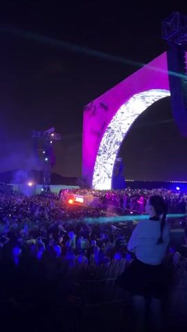 The video shows a large crowd of people at an outdoor concert at night. The stage is decorated with a large archway that is lit up in various colors, and there are lights shining across the crowd. A woman is standing near the camera, looking out at the crowd. There's a full moon visible in the sky. The event has vibrant lighting, creating an energetic atmosphere.