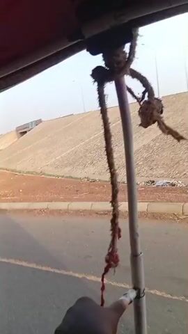 The video captures a first-person perspective from inside a moving tricycle in what appears to be Shuwarin, Nigeria. The driver's hand, holding a lit cigarette, rests on the handlebar in the foreground. Ropes are tied and hanging from the metal support beams of the tricycle's roof. Through the windshield, a clear, sunny daytime scene unfolds, revealing a road alongside a wide, sandy or dirt area. This area is populated with numerous makeshift market stalls or shelters constructed from poles and various coverings.

In the background, a large concrete embankment or bridge structure is visible, with streetlights positioned along its crest. Briefly at the start of the video, a person on a motorcycle can be seen ascending this embankment. The driver's reflection, wearing a light-colored shirt, is visible in the tricycle's side mirror. The sky is clear, indicating fair weather. The ambient sound includes the vehicle's engine and general environmental noise as the tricycle travels. No discernible text is visible in the frame.