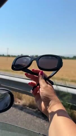 A person's hand with red nail polish holds up black sunglasses outside of a car window. The sunglasses are black and have tinted lenses. The background shows a bright blue sky and a field of dry grass. There is a metal guardrail and a side mirror of the car visible in the foreground. The video is shot in landscape orientation.