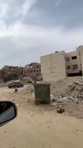The video shows a vehicle moving slowly along a dirt road in a desolate urban area. Buildings in varying states of disrepair line the road, with some showing clear signs of damage, like missing walls and exposed interiors. Piles of debris and rubble are scattered throughout the scene, and a few parked vehicles and a person walking can be seen. The sky is overcast, contributing to the somber atmosphere. The shot is taken from inside the moving vehicle, with the side mirror partially visible in the foreground.