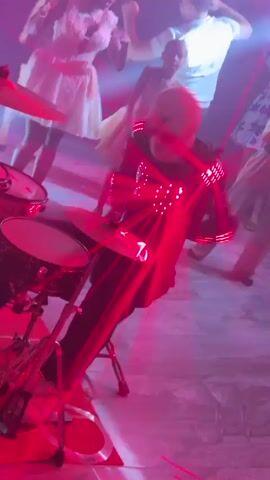 This is a low-angle, shaky video of a person playing drums at a party or club. The drummer is wearing a black outfit with red lights on their arms and chest, and a white mask. Red laser beams crisscross the scene, illuminated by the red lights on the drummer's outfit. Several other people are visible in the background, some dancing. The environment appears to be a dimly lit room with a dance floor. The video is shaky and the lighting is dynamic, making it difficult to clearly see the faces or actions of the background individuals.