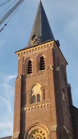 The video shows a close-up view of a brick church tower with a clock and a spire against a blue sky. To the left of the tower, a white object, possibly a crane or part of a carnival ride, is visible. The camera pans down to reveal the front entrance of the church, which has an arched doorway. Further to the left, a funfair ride with a trampoline-like surface and a colorful structure is prominent. Behind the ride, a white trailer with blue accents is parked. In the background, trees and more of the church building can be seen. The ground appears to be paved.