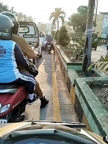 A POV shot from a motorcycle in traffic. There are several other motorcycles and a white pickup truck visible in front. The road has a yellow line dividing lanes. To the right of the road is a raised planter box with some plants. There is a palm tree next to the planter box. The sky is bright and looks like it is daytime.