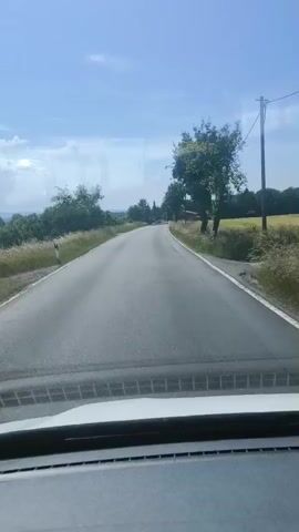 The media file captures a point-of-view perspective from inside a vehicle traveling along a rural road. The road is paved and appears to be a two-lane highway. The surrounding landscape is characterized by rolling green hills, lush vegetation, and scattered trees. In the distance, the terrain suggests a gently undulating countryside. The sky is predominantly blue with a few scattered white clouds, indicating fair weather and a clear day.

The scene is set during the daytime, likely late morning or early afternoon, given the brightness and the angle of the sun. The time of year appears to be summer or late spring, based on the vibrant green of the foliage. There are no people visible in the frame, and the focus is on the journey and the environment.

Notable details include road signs, which are partially visible on the side of the road. These signs are likely traffic or directional markers, typical of European roadways. The vehicle's interior is also visible in the foreground, including a portion of the dashboard and the windshield. A side mirror is also shown, reflecting a portion of the road and another vehicle behind.

The overall mood conveyed is one of peaceful travel through a scenic, natural setting. The movement of the vehicle suggests a journey or road trip. No text is clearly legible within the media file. The location context of Jena, Germany, implies the specific geographical area where this rural road is situated, suggesting a landscape typical of the Thuringian countryside.
