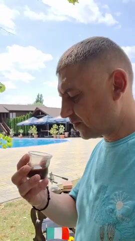The video shows a man in a turquoise t-shirt holding a small glass with a dark liquid, likely coffee, up to his lips and taking a sip. He has a close-cropped haircut and is outdoors, with a swimming pool and a building visible in the background. After drinking, he lowers the glass and smiles at the camera. A small table with some items on it, including a metal object, is to his left. The overall atmosphere appears relaxed and sunny.