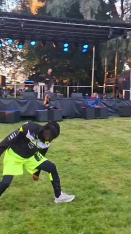 A man with braided hair and athletic wear, including a black and yellow jacket and neon green shorts with "BOSS" written on them, performs a dance move on a grassy field. He is facing away from a stage setup with lights and speakers. There are other people on and behind the stage, and trees are visible in the background. The scene appears to be outdoors during daylight.