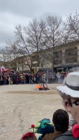 A public gathering appears to be in progress in a town square on an overcast day. A bonfire is in the center of the square, and a person dressed in an orange suit and equipped with a fire extinguisher is extinguishing the fire. Many people, including children dressed in costumes, are observing this event. Buildings and bare trees form the backdrop of the scene. A large mask representing a cartoonish cowboy is in the foreground.