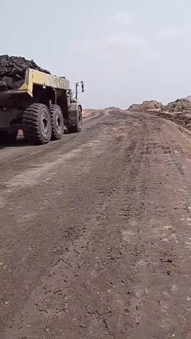 A large yellow dump truck filled with dark material, possibly coal, drives away from the camera on a dirt road. The road appears unpaved and muddy, with tire tracks visible. The background consists of a hazy sky and what looks like piles of dirt or hills on either side of the road. The truck gradually moves uphill.