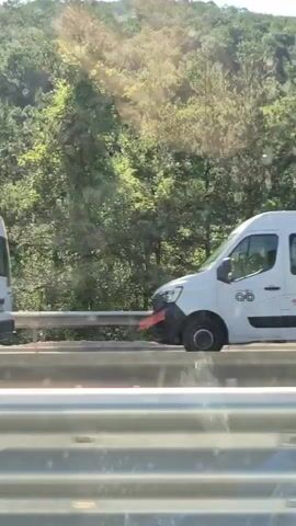 A white van with a logo on the side drives past a metal barrier. A yellow truck with a black hopper drives past the barrier. A yellow road roller with a black cab drives past the barrier. A wooded hillside is visible in the background.