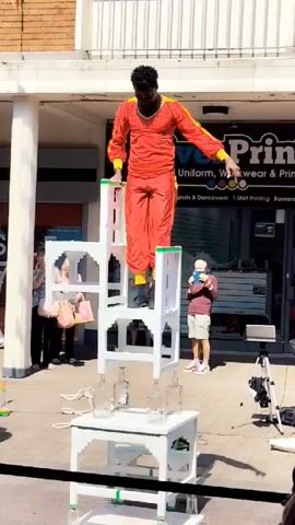 A man wearing a red and yellow jumpsuit stands on top of a stacked structure of white chairs, which are themselves balanced on top of a white table. He leans forward, performing a balancing act. The structure appears to be made of white plastic. Below the chairs, several glass bottles are placed on the table. In the background, a storefront with a sign that reads "Power Printing" is visible. Several onlookers are present, some taking pictures with their phones. The scene appears to be outdoors on a sidewalk.