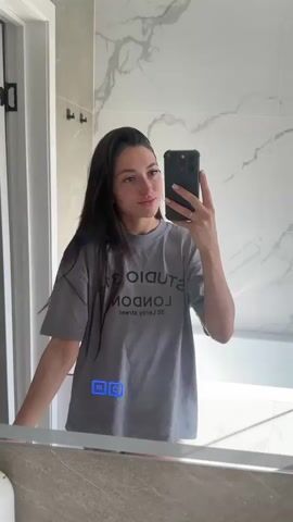 A young woman with long dark hair stands in front of a mirror, taking a selfie with her phone. She is wearing an oversized grey t-shirt with black text that reads "STUDIO 312 LONDON" and "38 LEROY STREET". The background appears to be a modern bathroom with marble tile walls and a bathtub visible. The woman is looking at her phone and smiling slightly. There are some illuminated icons on the mirror in the lower left corner of the frame.