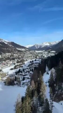 The video shows a scenic view of a town nestled in a valley among snow-covered mountains, captured from what appears to be a moving cable car. The town is a mix of residential and commercial buildings, with snow-covered roofs and some open, snow-covered spaces. The surrounding landscape is a mix of evergreen trees and snow-covered slopes. The sky above is a clear, bright blue. The angle of the shot suggests a high vantage point, with the cable car wires visible at the top of the frame.