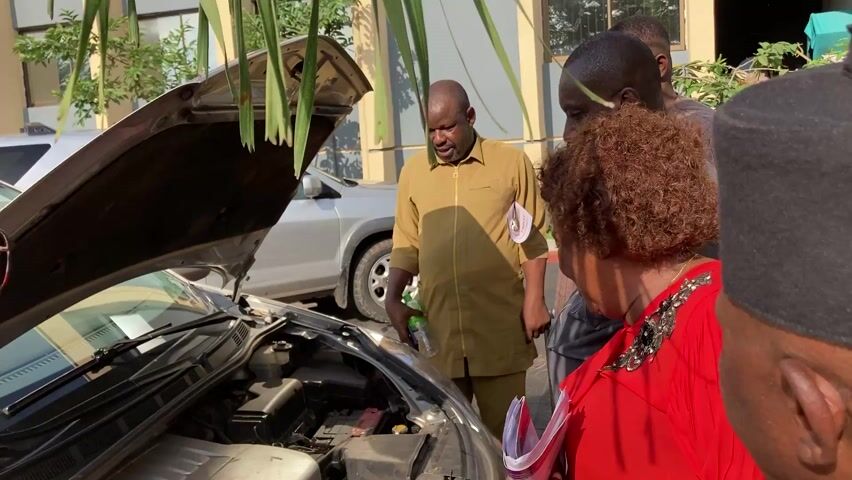 Four people are standing around a car with its hood open, looking at the engine. The car is silver, and another silver vehicle is parked behind it. The person closest to the camera is a woman in a red top. To her right is a man in a tan shirt and pants. Behind him is another man in a striped shirt. The fourth person is partially visible, with a black beanie on their head. They are standing to the left of the car. The background includes a building and some greenery. The lighting suggests it is daytime.