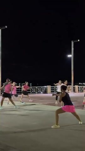 A group of six women are doing a form of aerobic exercise in an outdoor paved area at night. They are all wearing athletic clothing, mostly pink tops and skirts. The women are spaced out and appear to be following a leader who is not fully visible. The environment is dark with some artificial lighting from poles. There is a railing and a body of water in the background.