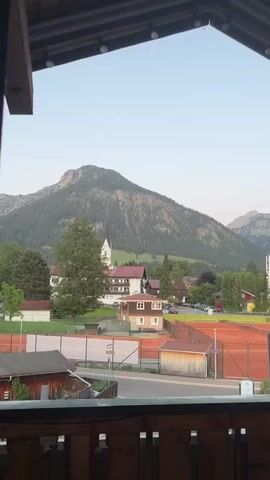 The video shows a view from a balcony overlooking a picturesque alpine village. In the distance, a large, green mountain dominates the landscape, with a smaller mountain range visible to the right. The village below features a church with a tall steeple, several buildings with red roofs, and lush green trees. In the foreground, there are orange tennis courts and a small wooden shed. A road separates the tennis courts from the balcony. The sky is light blue, and there's a faint contrail from an airplane visible. The balcony has a wooden railing and roof, framing the scene. There are no people visible. The overall impression is of a serene, peaceful setting.