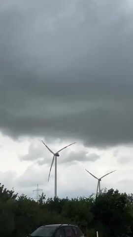 The video shows a view from a vehicle driving on a highway, looking at a cloudy sky and wind turbines. The turbines have white towers and red tips on their blades.  Green trees are visible below the turbines. A blue highway sign indicating a rest area ("Rastplatz") with an arrow pointing to the right is visible as the vehicle moves forward.