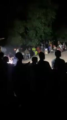 At night, a dimly lit open-air gathering shows a large group of people spread across a dusty or sandy area. Silhouetted figures stand in the foreground, appearing to observe the central activity. Some people are seated in the center of the space, possibly watching a performance or participating in an event. Trees surround the edge of the area, obscuring the background further and creating a sense of enclosure. The lighting is minimal, making it difficult to distinguish specific details and features of individuals or objects.
