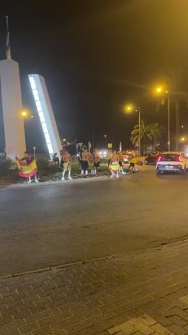 A group of people, some of whom are wearing minimal clothing, stand on a circular roadside area, waving Spanish flags. Several cars, including a red one, pass by them. In the background, there is a large white structure that appears to be a monument or sculpture, illuminated by lights. The scene takes place at night, with streetlights also visible. The road surface is wet, suggesting recent rain.