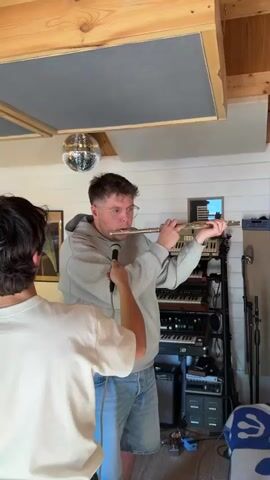 This video captures two men in a well-lit music studio or rehearsal space. The room features white walls, wooden ceiling beams, and grey acoustic panels on the ceiling, suggesting attention to sound treatment. A reflective disco ball hangs from the ceiling, adding a decorative element.

One man, seen from behind in a white t-shirt, holds a black microphone directed towards the other man. The second man, wearing a grey hoodie, blue denim shorts, and clear-rimmed glasses, is actively playing a silver flute. He appears focused on his performance, with his mouth at the flute's embouchure hole and his fingers pressing the keys. The microphone's position indicates that the flute playing is being recorded.

To the right of the flutist, a multi-tiered stand holds a stack of electronic keyboards, synthesizers, and other audio equipment. A speaker or amplifier unit is visible on the floor beneath this equipment. The scene depicts a collaborative recording session, likely for a musical project, in what appears to be a casual yet equipped studio environment located in the Borough of Runnymede, United Kingdom. No discernible text is visible in the frame.