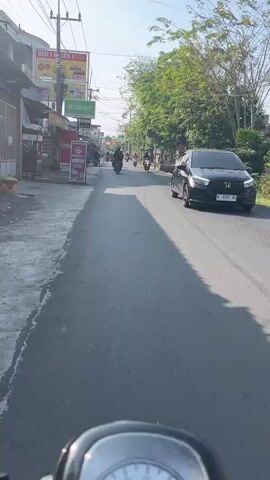 The video is a first-person perspective shot from a scooter on a paved road. Several other scooters and a car are visible in the distance, moving in the same direction. The road is lined with shops and trees on both sides. The sun is shining, casting shadows on the road. A speedometer is visible at the bottom of the frame.