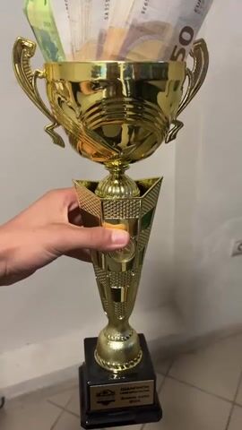 A person's hand holds a golden trophy, rotating it to show its details and contents. The trophy is intricately designed with a shiny golden finish and rests on a black square base. Embedded in the middle section of the trophy's stem is a circular gold medal-like ornament. The base features a small plaque with text in Cyrillic, which reads "ЧЕМПИОН" (Champion) and "Зима 2020" (Winter 2020), along with some other partially visible words that appear to be related to a sporting event or stream (e.g., "СПОРТНО ПОТОК").

The cup part of the trophy is filled with numerous Euro banknotes. Visible denominations include 100 Euro notes (green) and 50 Euro notes (orange). The banknotes are fanned out, showcasing their colors and the distinct Euro currency markings. Some visible serial numbers and "EBC 2017" are also present on the notes.

The setting appears to be indoors, with a plain light-colored wall and a tiled floor visible in the background. The lighting is consistent with an indoor environment. The hand holding the trophy is of a light skin tone. The video focuses closely on the trophy and the money inside it, emphasizing the reward aspect.