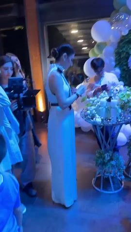 The video opens on a dimly lit indoor event space, possibly a party or reception. A woman in a long white dress with a black collar and belt is the primary focus. She stands facing a table laden with white candles and floral arrangements, holding a small box in her hands and looking down at it. To her left, a woman in a black dress and a woman in a white top are partially visible, engaged in conversation. Behind the main subject, a person wearing glasses and a white shirt is seen looking at their phone, near the floral display. The background features a wall adorned with numerous white and green balloons, and further back, a statue of an angel is visible. The lighting is a mix of warm overhead lights and cooler, greenish spotlights, creating a somewhat theatrical atmosphere. The scene transitions between slight camera movements, offering different perspectives of the individuals and the decor.