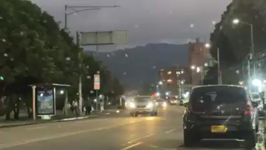 The video shows a street scene on a rainy evening. Several cars are driving on the road, with a bus stop on the left side of the frame. A cyclist wearing a bright pink delivery backpack with the Rappi logo rides in front of the cars. Buildings and mountains are visible in the background.