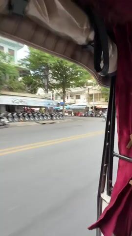 This is a first-person perspective video, likely from inside a tuk-tuk. The camera is positioned to look out the side and slightly forward. A street is visible with other vehicles, including motorcycles and cars. There are buildings and trees lining the street. Several motorcycles are parked on the side of the road. The interior of the tuk-tuk is partially visible, with red curtains and a tan headliner. The video is in motion, indicating the tuk-tuk is traveling. The time stamp shows the video is approximately 5 seconds long.