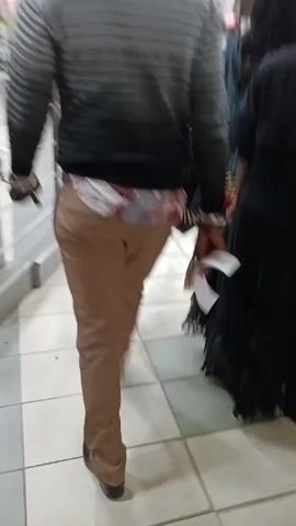 The video shows a person walking through a shopping mall. The person is wearing a grey and black striped sweater, brown pants, and black shoes. They are carrying a phone in their hand. The camera follows the person from behind as they walk past various shops and food vendors. Other people are visible in the mall, some walking and others seated at tables. There are signs for "Creamy Inn" and "Pizza Inn" visible in the background. The mall has a tiled floor and bright overhead lighting.