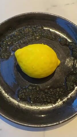 A close-up, overhead shot shows a bright yellow lemon on a dark, textured plate. The plate is rimmed with a dark, gelatinous substance that appears to be caviar or boba. A silver spoon is used to scoop out a portion of the lemon's flesh. The lemon is partially peeled, revealing the white pith underneath. The background is blurred, suggesting a kitchen counter or table.