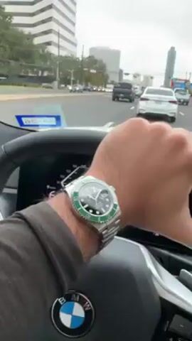 A person is driving a white BMW through a city on an overcast day. Traffic is moderate. The driver is wearing a Rolex watch. The vehicle has a touchscreen that shows Apple Carplay with a map and music. Buildings and traffic signals line the street. The view is through the windshield of the car.