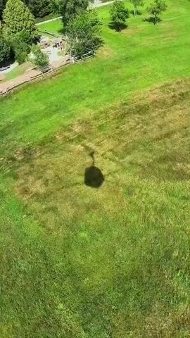 This is a drone shot looking down at a grassy field with a shadow of a gondola cast on it. The gondola appears to be moving from right to left. In the upper left corner of the frame, there is a playground area with some structures and trees. The grass is green and well-maintained. The sky is not visible in the frame.