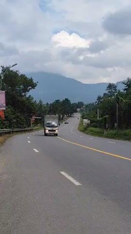 A truck drives down a winding road surrounded by trees and mountains. The sky is cloudy. The truck is white and has a metal cargo box. There is a yellow line down the middle of the road. The video is shot from a slightly elevated angle, looking down the road.