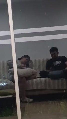 A video shows two people sitting on a couch. The person on the left is a woman with dark hair, wearing a light-colored hoodie and jeans. She is holding a white pillow and appears to be resting her head on it. The person on the right is a man with dark hair and a beard, wearing a black t-shirt and jeans. He is holding a black object, possibly a phone, in his right hand. Both individuals appear to be relaxed and are looking down, perhaps at their phones or the object in the man's hand. The couch is striped with beige and white. Behind them, a wall is visible with horizontal white stripes. The environment appears to be outdoors, with some greenery visible in the foreground, suggesting it might be a patio or backyard. The lighting is dim, indicating it might be evening or nighttime.