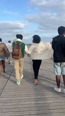 A group of people walks along a wooden boardwalk. In the foreground, a person with a brown backpack walks behind a person wearing a white top and black pants. Another person walks on the left side of the frame, and a person on the right side of the frame talks on the phone. The group is walking toward the distant horizon where there is a bridge and a body of water. The sky is partly cloudy.