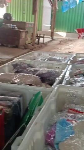The video shows an outdoor market stall with rows of plastic crates filled with packaged clothing. The crates are white and the clothing inside is wrapped in plastic. Some of the packages have visible labels with text like "SIMPLE FIT". In the background, there is a green corrugated metal wall, a wooden structure, and a red wheelbarrow. The ground is dirt. There are some people in the background but their faces are not visible. The lighting suggests it is daytime.