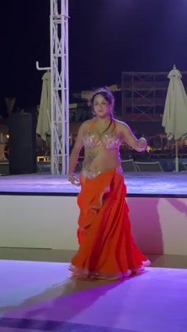 A woman is performing a belly dance on an outdoor stage at night. She is wearing a sparkly bra top, an orange skirt with frills, and jewelry. The stage is illuminated with pink and purple lights. Behind the stage, there is a swimming pool area with sun umbrellas and a water slide. At one point a child walks across the foreground.