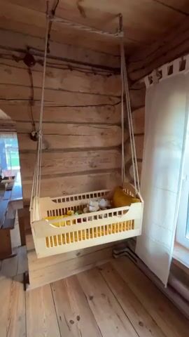 A wooden baby crib is suspended from the ceiling by ropes. The crib is filled with stuffed animals and blankets. The crib is rocking gently. The room is made of wood and has a window with white curtains.