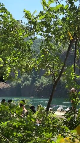 The video shows a scenic outdoor location with lush green foliage in the foreground, obscuring a clear view of a body of water with several people. The water appears to be a lake or quarry, with steep, rocky cliffs in the background. Sunlight filters through the trees, creating dappled shadows. In the water, several individuals are visible, some swimming and others on what appear to be inflatable rafts or paddleboards. Along the edge of the water, closer to the camera, a group of people are gathered, sitting and interacting. One person is standing near the edge of the water. The overall atmosphere suggests a recreational setting on a bright, sunny day.
