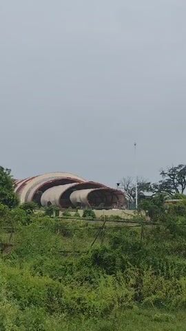 The video shows an exterior view of a large, modern, partially constructed or possibly abandoned building with a unique, curved, shell-like architectural design. The structure is nestled amongst lush green vegetation and trees. The sky above is overcast and hazy. The camera pans slowly across the building, revealing its multi-layered, swooping rooflines and open sections. Fencing is visible in the foreground, and some debris or overgrown areas suggest the site might be undeveloped or neglected. No people or animals are visible. The overall impression is one of a striking but possibly unfinished or disused architectural project surrounded by nature.