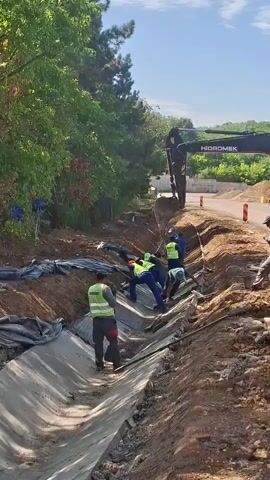 A construction crew is working on a drainage ditch. Several workers in high-visibility vests are in the ditch, some using shovels and others appearing to be adjusting or placing material along the sides of the ditch. An excavator is visible in the background, its arm extended over the ditch. The environment appears to be outdoors, with trees and vegetation on the left and a road or cleared area on the right. The sky is clear and blue. The ditch itself has a concrete or similar lining.