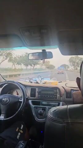 The video shows the interior of a car as it drives on a highway. The camera is positioned behind the driver, looking forward. The dashboard is visible, including the steering wheel, radio, and air conditioning controls. A person's hands are on the steering wheel. Outside the car, trees and other vehicles are visible. The sky is bright, suggesting it is daytime. The road appears to be in good condition, and there are safety barriers on the side of the road. The overall impression is one of a normal car journey on a highway.