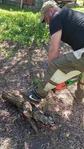 A man stands outside using a chainsaw to cut a fallen tree branch. He wears a black t-shirt, tan pants with white patches, a tan baseball cap, and sneakers. The man has safety gloves on his hands. He places his left foot on the branch to hold it in place. He uses the chainsaw to cut the branch into smaller pieces. The area around the man is grassy with some small plants growing. There is a small wooden structure in the background. The weather is sunny. Once finished cutting, the man lifts the chainsaw and moves it away from the cut branch.