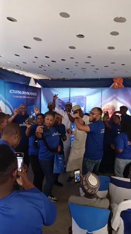A group of people, mostly dressed in matching blue t-shirts with the text "Grow Active Grow Ready" on the back, are gathered in a room. They are standing, dancing, and clapping, with many holding up their phones to take pictures or videos. A man in a white traditional robe and hat is at the center of the group. There's a backdrop with text that reads "ACTIVATING GROWING MOMENTS" and a logo with the word "LACTOGROW." The room has a white ceiling with circular lights. In the foreground, another person is seated, also holding up a phone to record the event. Overall, the scene depicts a celebratory gathering, possibly a product launch or promotional event.
