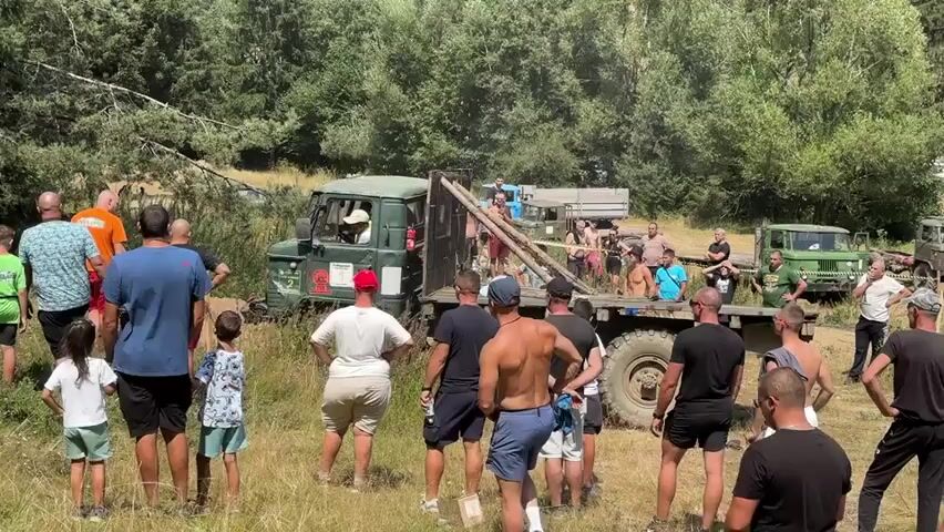 A group of people, mostly men and some children, are gathered in a wooded area on a sunny day. They are watching a green truck with a flatbed trailer that appears to be stuck in the mud. Some people are standing near the truck, while others are observing from a distance. One person is sitting in the driver's seat of the truck. There are other vehicles visible in the background, including another truck and a trailer. The ground is covered in dry grass and there are trees and brush in the background.