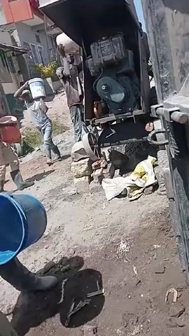 The video depicts a construction site with several individuals engaged in manual labor. A worker wearing rubber boots and gloves is seen inserting their arms into a large, rusty metal barrel, seemingly to retrieve or mix something inside. Adjacent to the metal barrel is a blue plastic container and a white pipe lying on the ground. In the background, other workers are seen carrying bags or buckets, and a small machine with an exposed engine is visible. The ground is uneven and covered with dirt, mud, and scattered debris, indicative of an active construction environment.