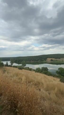A video shows a landscape with a lake, trees, and fields. The sky is cloudy. The camera pans from right to left, revealing a body of water with reflections of trees and sky. In the foreground, there is dry, tall grass. Beyond the lake, there are green trees and agricultural fields, with rolling hills in the distance. The overall impression is of a rural and natural setting.