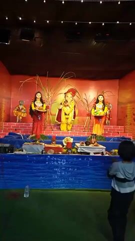 The video shows a shrine with religious statues. In the center is a statue of the Hindu deity Ganesha, dressed in a yellow and maroon outfit with a red turban and a garland of yellow flowers. To his left and right are statues of women, also dressed in colorful attire and adorned with jewelry. The women hold offerings, one with a tray of what appears to be food, and the other with a basket of fruits. The statues are placed on a multi-tiered altar decorated with blue fabric. In front of the altar, various offerings like flowers and food items are arranged. Tall, dry grass stalks stand behind the central Ganesha statue. A young child, facing away from the camera, stands in the foreground, observing the shrine. The background appears to be a dark stage area with decorative lighting. The overall atmosphere suggests a religious ceremony or festival.