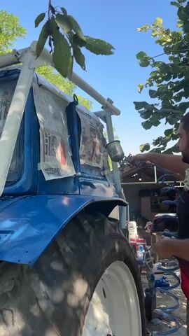 A man with a beard is spray painting a blue tractor. He is wearing a black t-shirt and red shorts. He is holding a spray gun in his right hand and a can of paint in his left hand. The tractor is parked outdoors in front of a building. There are trees and a blue sky in the background. The tractor is partially covered with newspapers to protect it from the paint. The man is standing next to the tractor and is focused on his work. He is wearing a mask to protect himself from the paint fumes.